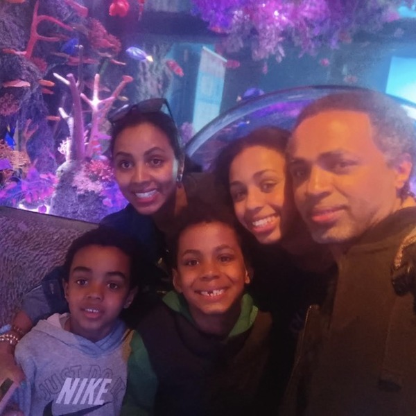 Five people gather closely for a group photo inside a colorful aquarium-like setting. Branching coral forms and saturated purple, pink, and blue lighting fill the space behind them. The figures are arranged in two rows, with faces turned toward the camera.