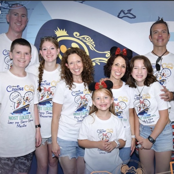 A group of people wearing matching white graphic shirts pose together in front of a decorated display. Several figures stand in the back row while others cluster across the center and front. Blue, white, and dark graphic elements fill the event-style background.