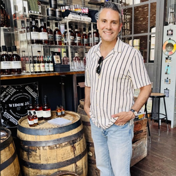 A person stands beside a wooden barrel inside a shop lined with bottles and merchandise. Shelving, industrial windows, and warm wood surfaces fill the interior. Daylight from the windows combines with indoor lighting across the display area.