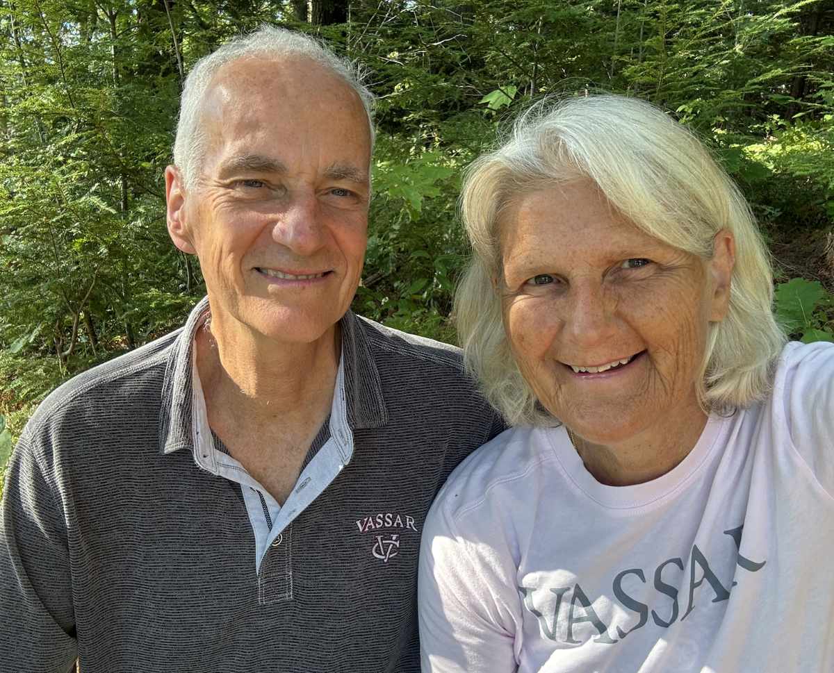 Two people pose closely together for an outdoor selfie in a wooded setting. Their faces fill much of the frame, with leafy green vegetation extending behind them. Soft daylight filters through the trees and creates gentle, even illumination.