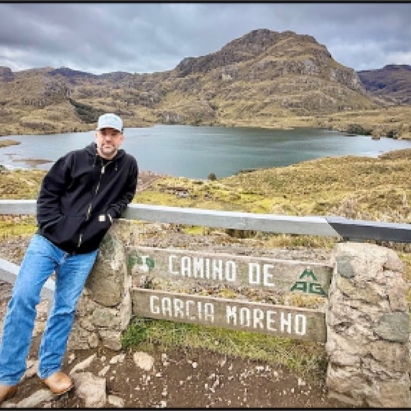 A person stands beside a stone viewpoint sign reading “Camino de García Moreno.” A mountain lake and rugged brown-green peaks spread across the landscape behind the figure. Diffused daylight falls beneath a cloudy gray sky.