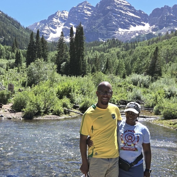 Two people stand beside a shallow mountain stream, with one figure slightly behind the other. Dense green shrubs and tall evergreens fill the valley around them. Snow-streaked rocky peaks rise sharply beneath a clear blue sky.