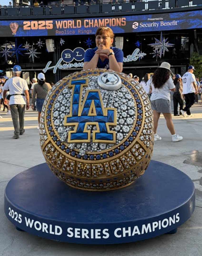 A person stands behind a large jeweled LA championship ring display at a stadium. The circular display reads “2025 World Series Champions,” with crowds and seating visible beyond it. Bright daylight illuminates the metallic blue, gold, and silver details.