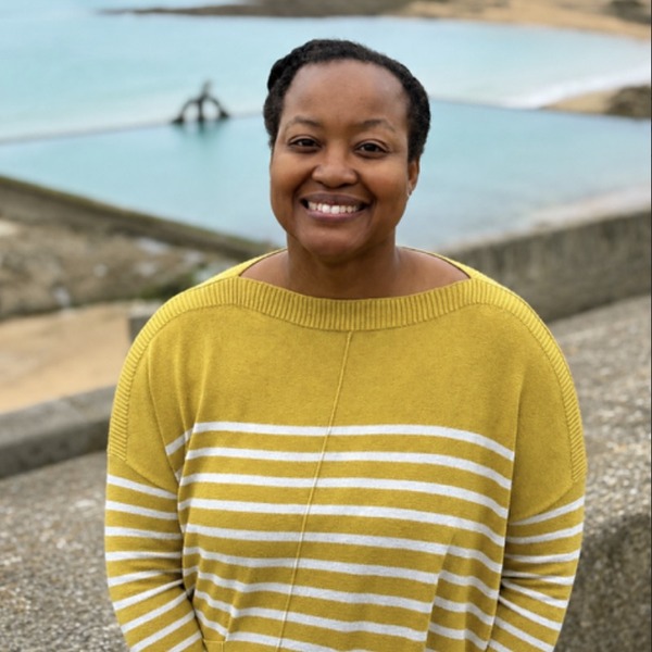 A person in a mustard-yellow striped sweater stands near a coastal overlook. Pale turquoise water, sandy shoreline, rocky outcrops, and green headlands extend across the background. Soft daylight falls under a lightly clouded blue sky.