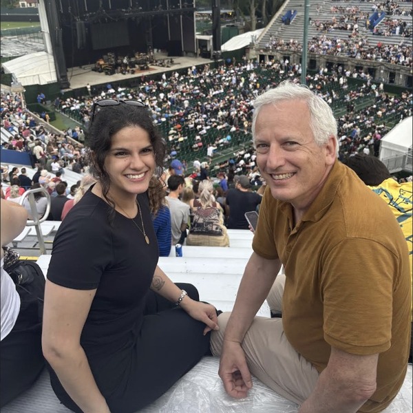 Two people sit together in stadium-style seating at a large outdoor concert venue. A broad stage, lighting structures, and a dense crowd fill the area behind them. Daylight and an overcast sky create even illumination across the scene.