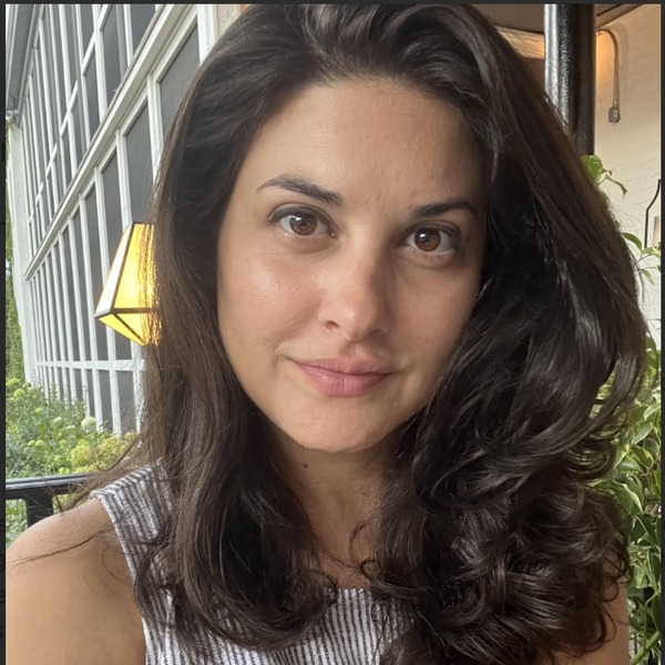 A close portrait shows a person with dark wavy hair wearing a sleeveless striped top. The figure faces the camera beside a windowed wall, with part of an indoor room visible behind. Soft natural light highlights the face and hair.