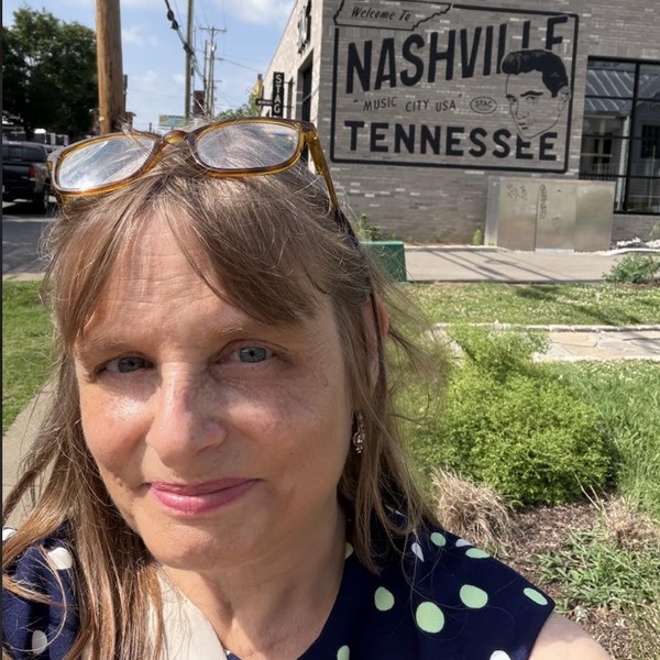 A close portrait shows a person outdoors with sunglasses resting on top of the head. Behind the figure, a weathered building bears large painted words reading “Nashville Tennessee.” Green grass, utility lines, and a bright blue sky with scattered clouds surround the structure.