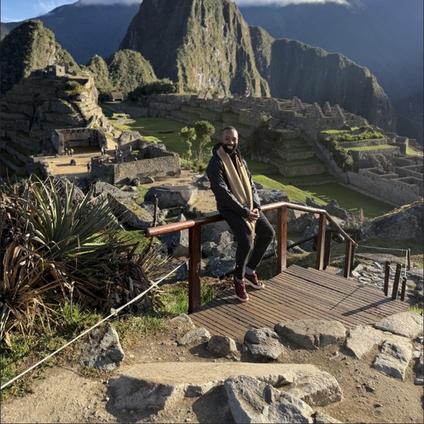 A person stands on a stone terrace overlooking Machu Picchu. Layered stone ruins, green terraces, and steep mountain peaks fill the valley below. Strong sunlight creates crisp shadows beneath a vivid blue sky with white clouds.