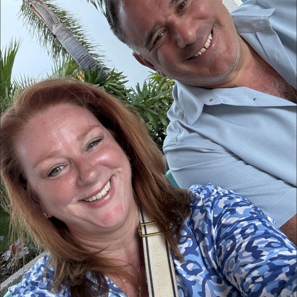 Two people pose closely together outdoors near palm trees and a turquoise-and-white building. One figure is positioned lower in the frame while the other leans in from above. Bright daylight creates clear highlights across their faces and clothing.