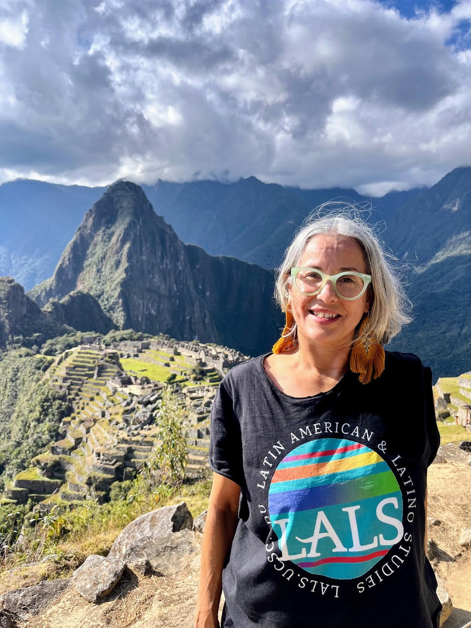 Catherine Ramírez smiling outdoors with mountains and ancient stone ruins behind her.
