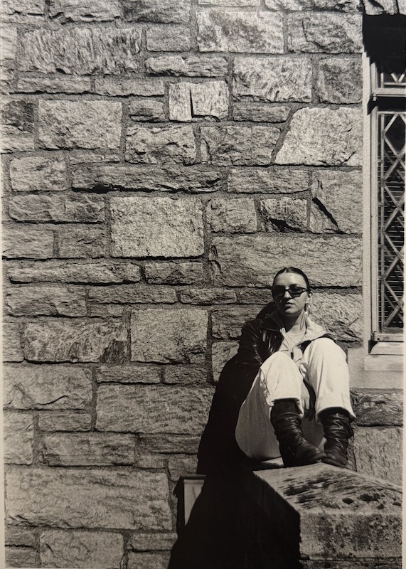 An artistic sepia-toned image of a person wearing sunglasses sitting in front of a stone wall.