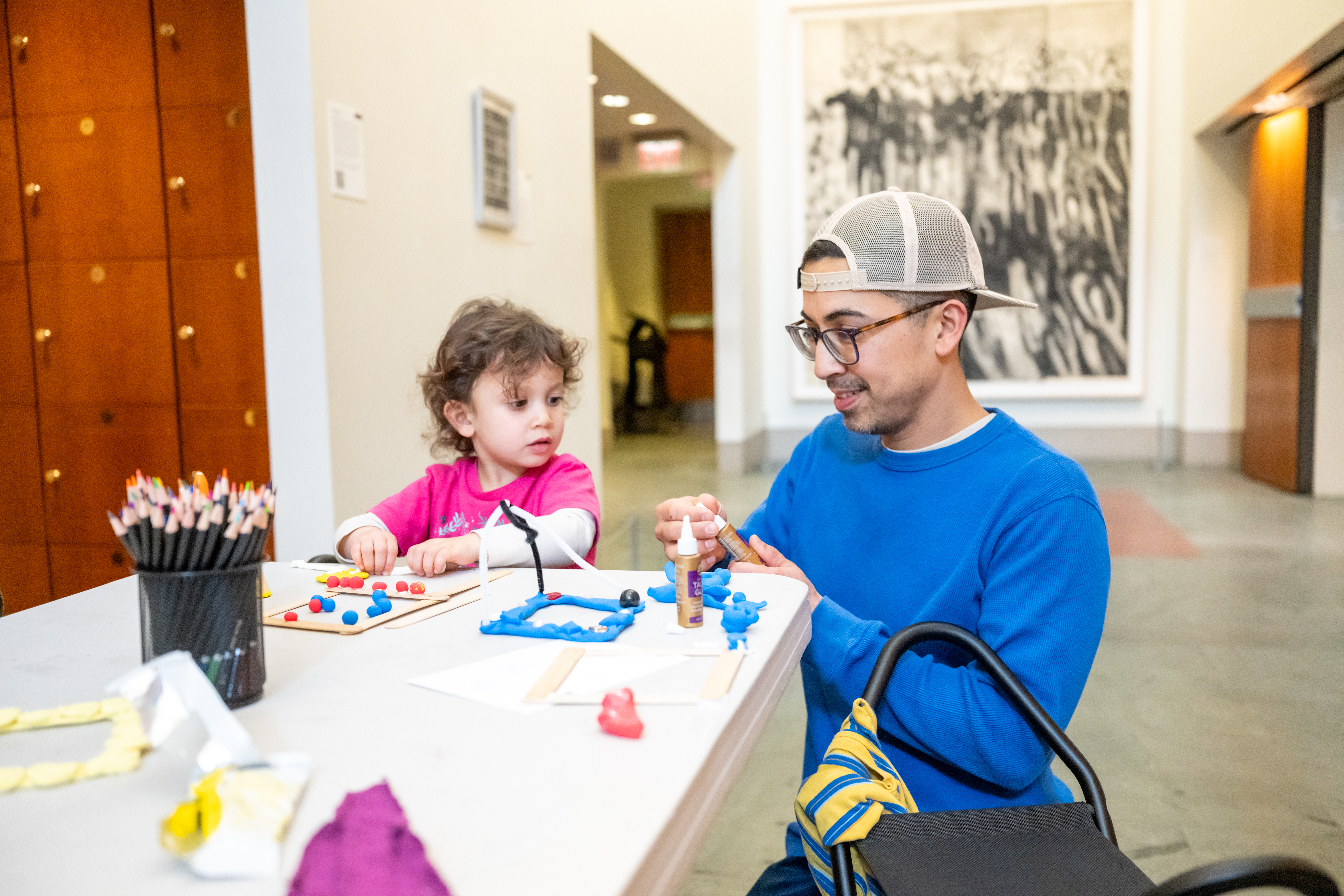 Photo of a man and young girl seated at a table making art with clay, colored pencils, and glue.