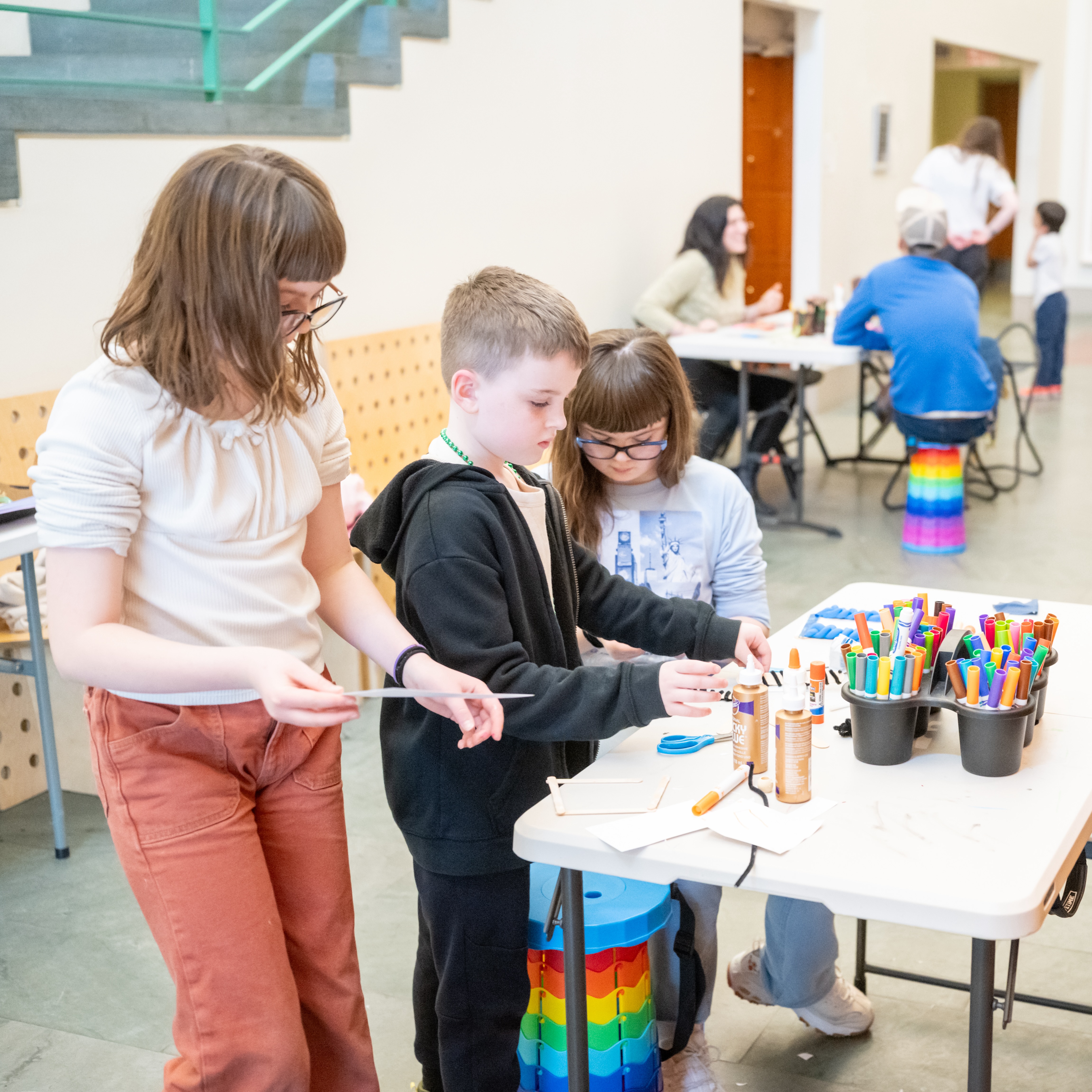 Photo of two girls and a boy making art around a table with clay, glue, markers, and scissors. 