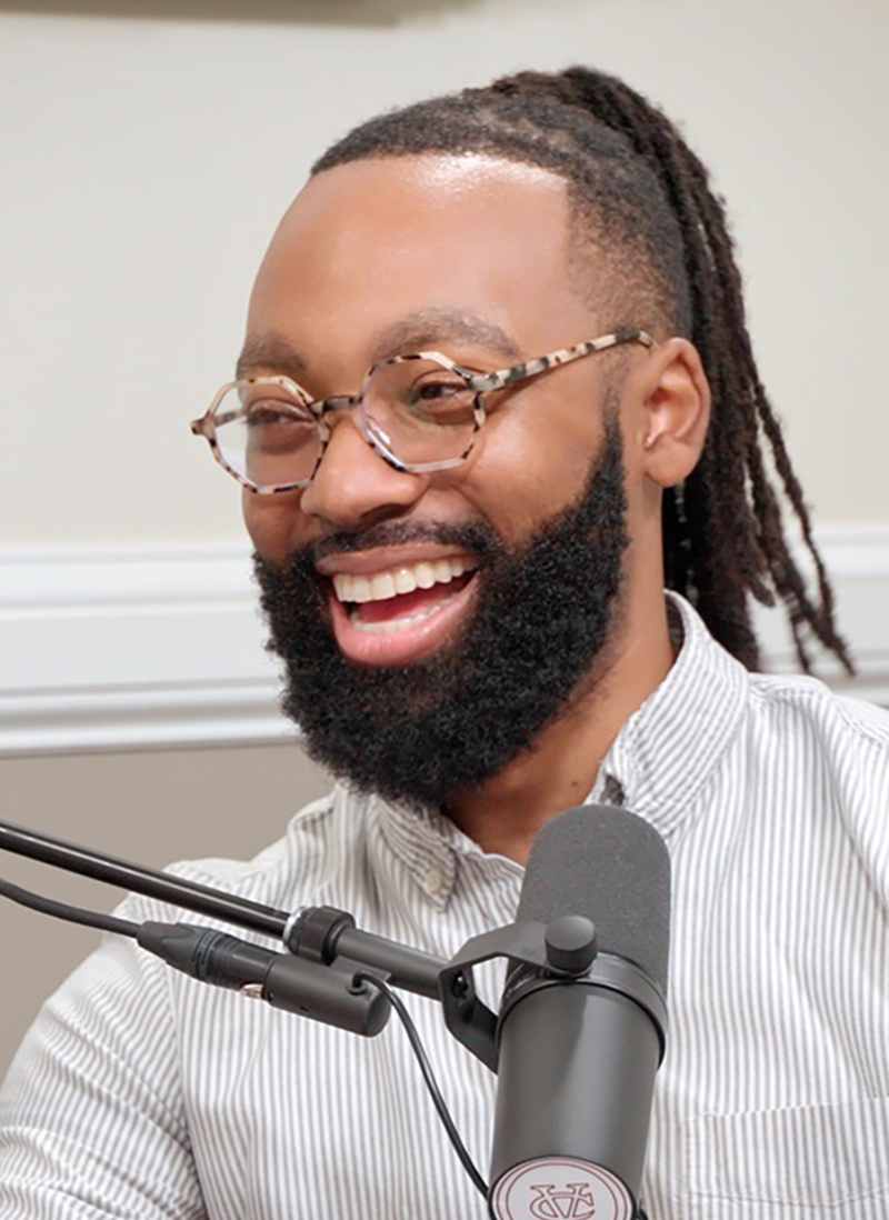 A portrait of Wesley Dixon, a person with dark braided hair, a dark beard, and a white shirt.