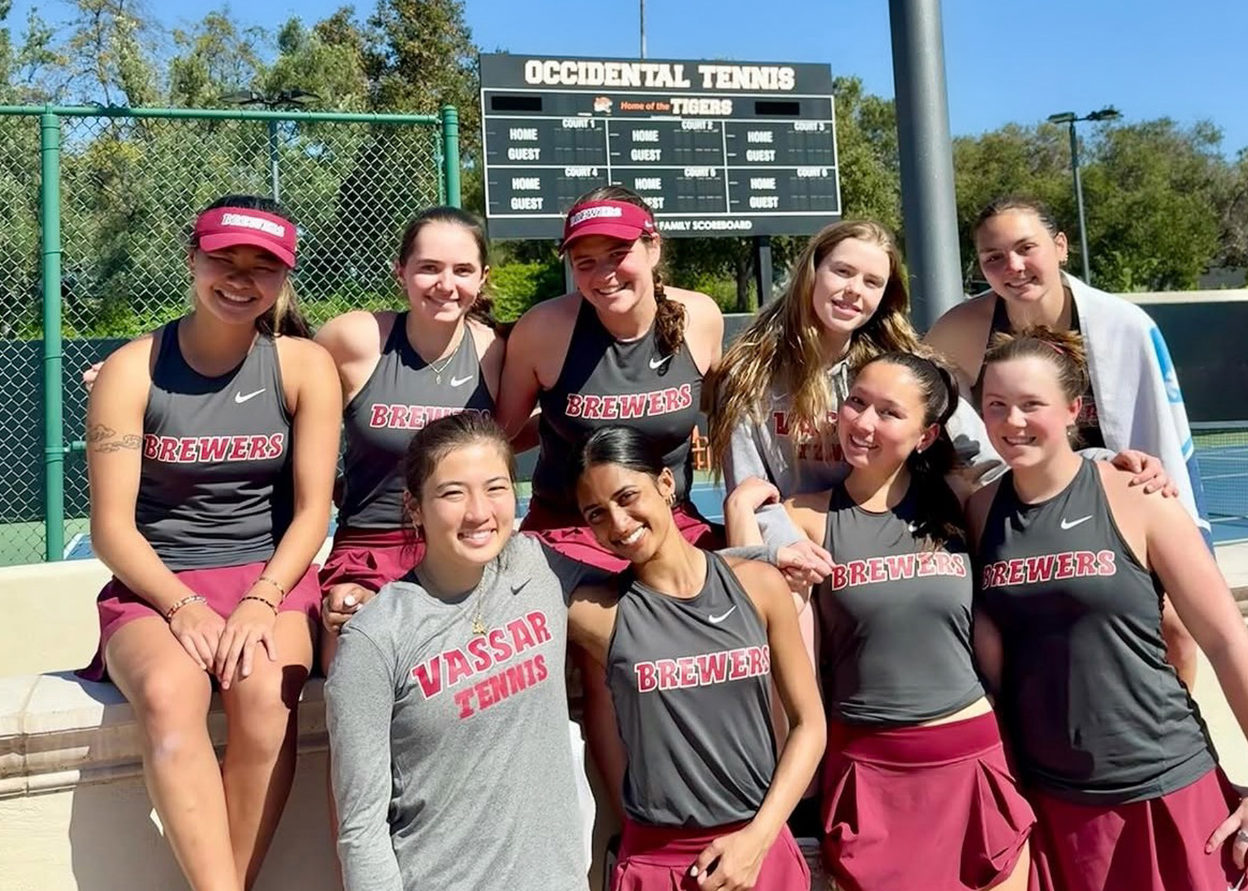 Eight tennis players in gray and burgundy Vassar uniforms pose together beside an outdoor tennis court.
