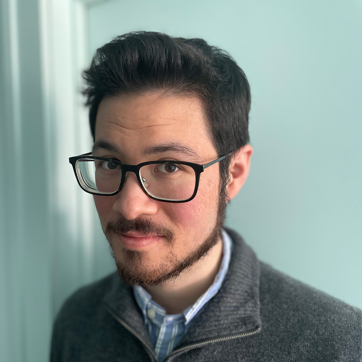 Luke Soucy, with dark hair, a neat beard, and black-framed glasses, wearing a grey quarter-zip sweater over a blue collared shirt against a soft teal background.