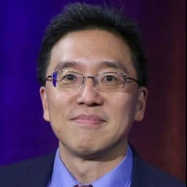 Charles Tien wearing wire-rimmed spectacles and has short black hair and a slight smile, dressed in a dark suit jacket over a light blue collared shirt. The background is a solid, dark purple-blue color.