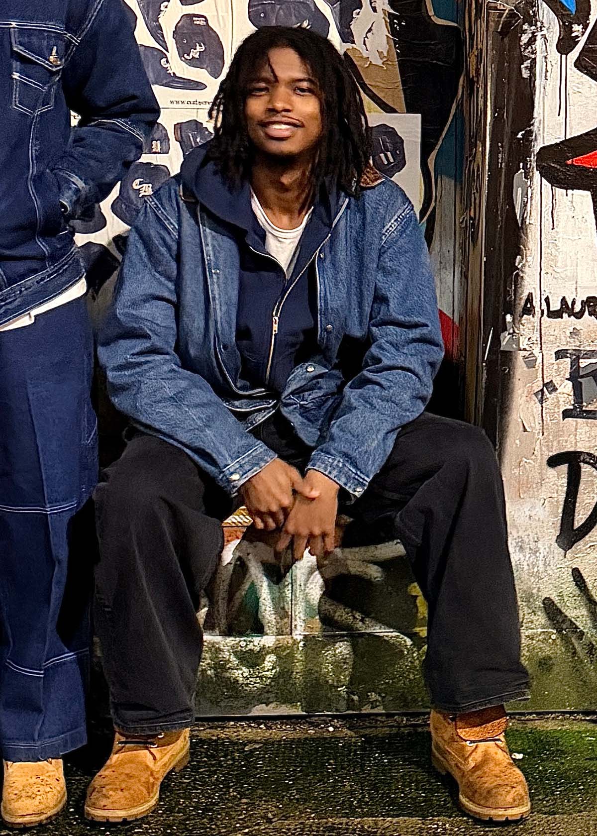 Gene Waddy with shoulder-length dark hair, sits outdoors against a wall covered with graffiti art. Gene is layered in a blue denim jacket over a navy zip-up hoodie, dark trousers, and worn tan work boots, leaning forward with his hands clasped between his knees.