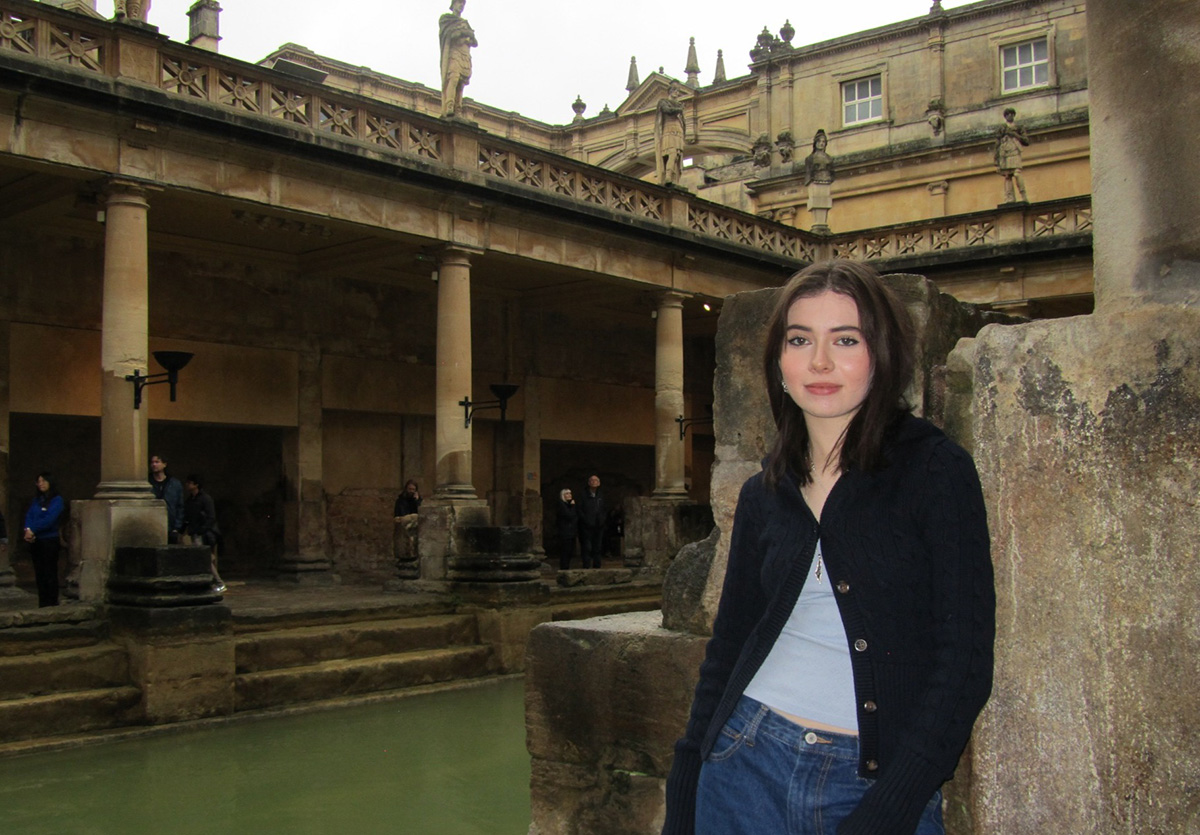 Isabella Zine wearing a navy cardigan and jeans leaning against a stone pillar inside the ancient Roman Baths, with stone columns, statues, and thermal waters in the background.