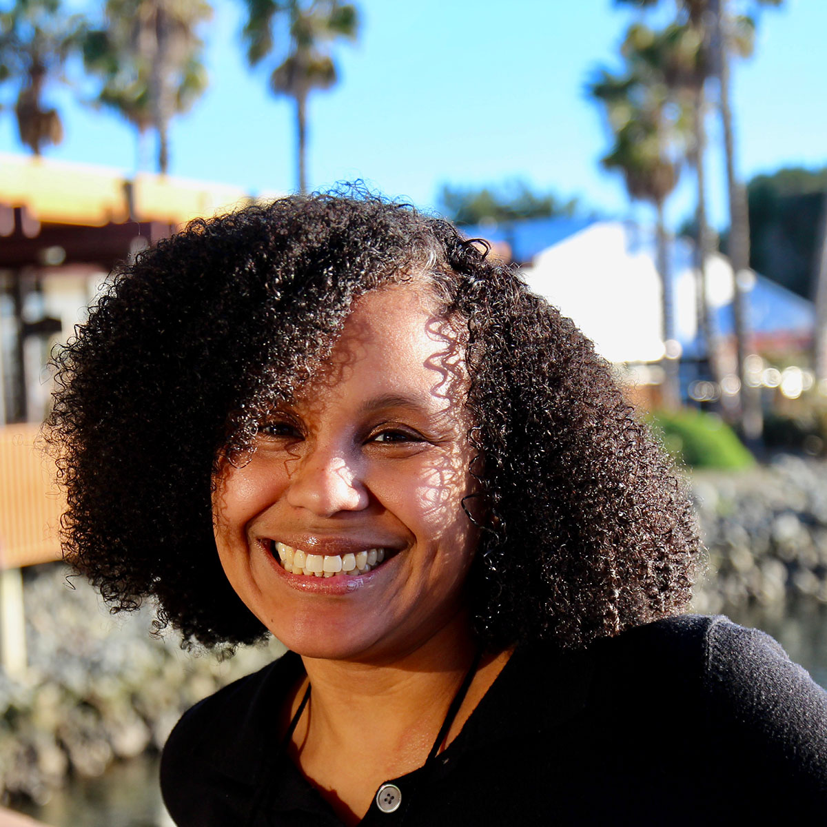 Katie Acosta with dark, shoulder-length curly hair smiling brightly in sunlight, wearing a black top against a blurred background of palm trees.