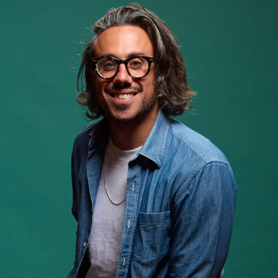 Nicholas Occhiuto smiling, with shoulder-length wavy hair, tortoiseshell glasses, and light facial hair, wearing an open denim shirt over a white t-shirt against a solid teal background.