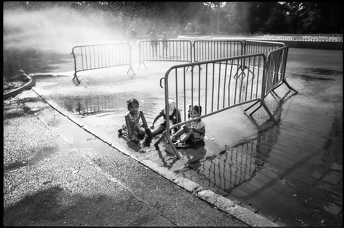 A black-and-white photograph taken by the artist Jas Leon. In the picture are three children playing in a spray of water while sitting in a city street. &nbsp;&nbsp;