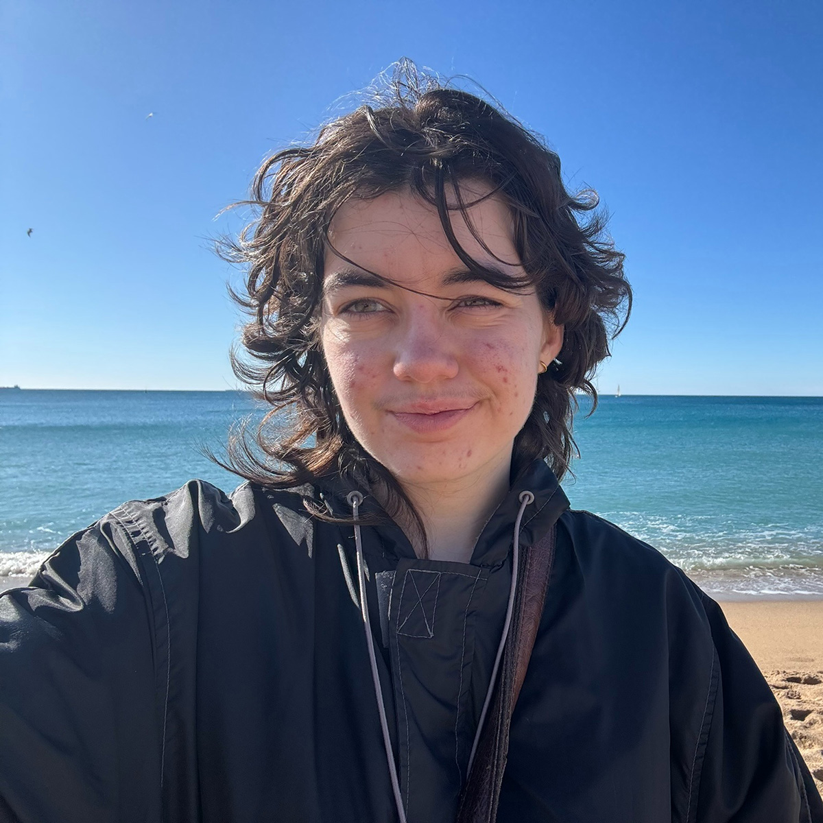 Sam Evans with dark hair, wearing a black jacket on a sunny beach with the blue ocean and clear sky in the background.
