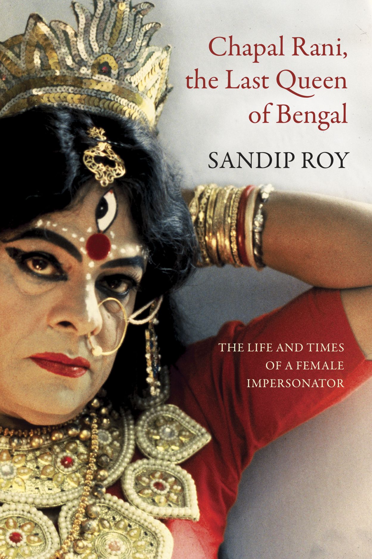 Book cover titled "Chapal Rani, the Last Queen of Bengal" by Sandip Roy. A person wears elaborate gold jewelry, a crown, red clothing, and theatrical facial makeup with a third eye, resting an arm stacked with bangles near their head.