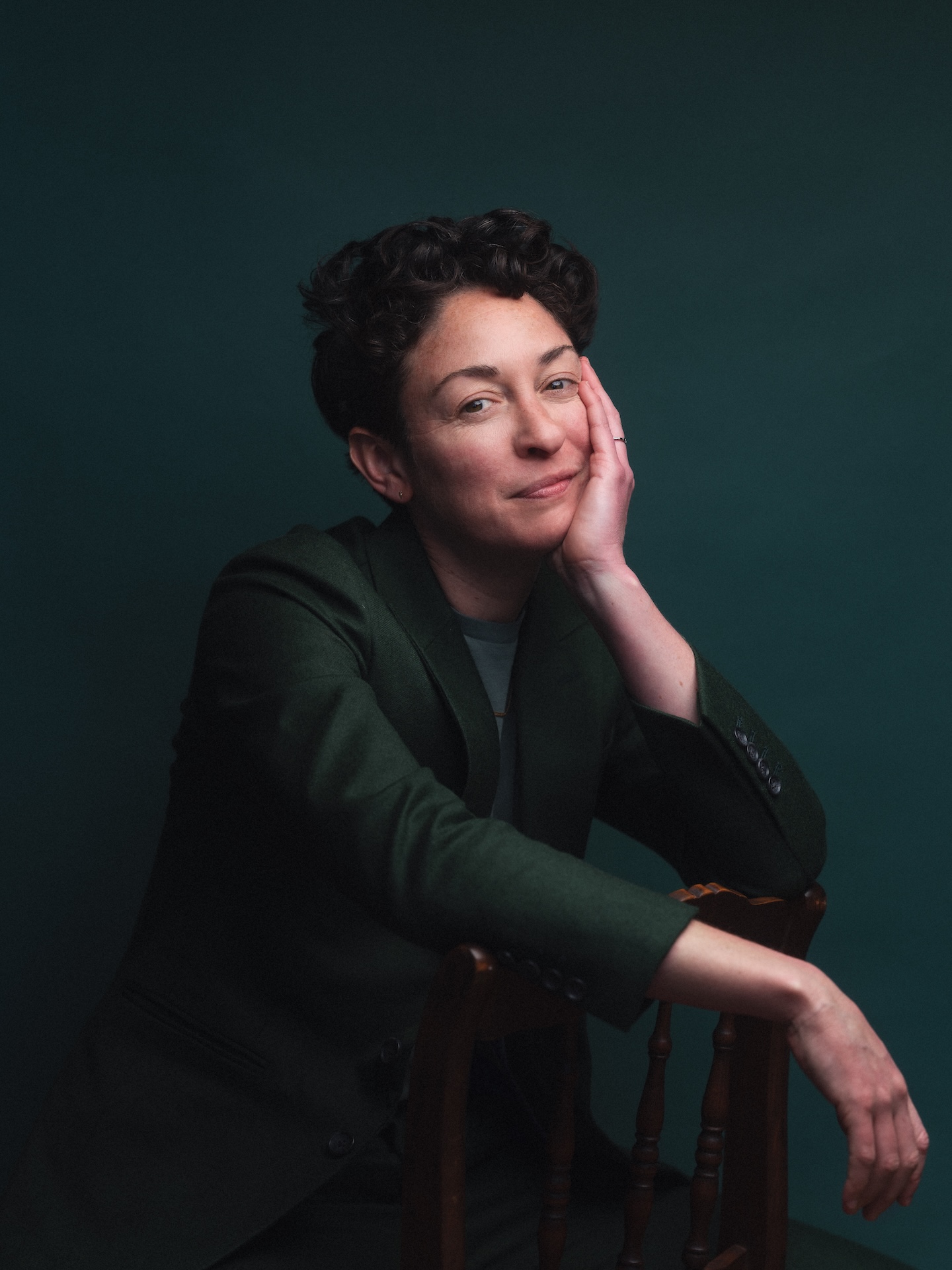 A person with short, dark curly hair wearing a dark green jacket rests their cheek against one hand while leaning over the back of a wooden chair against a solid dark green backdrop.