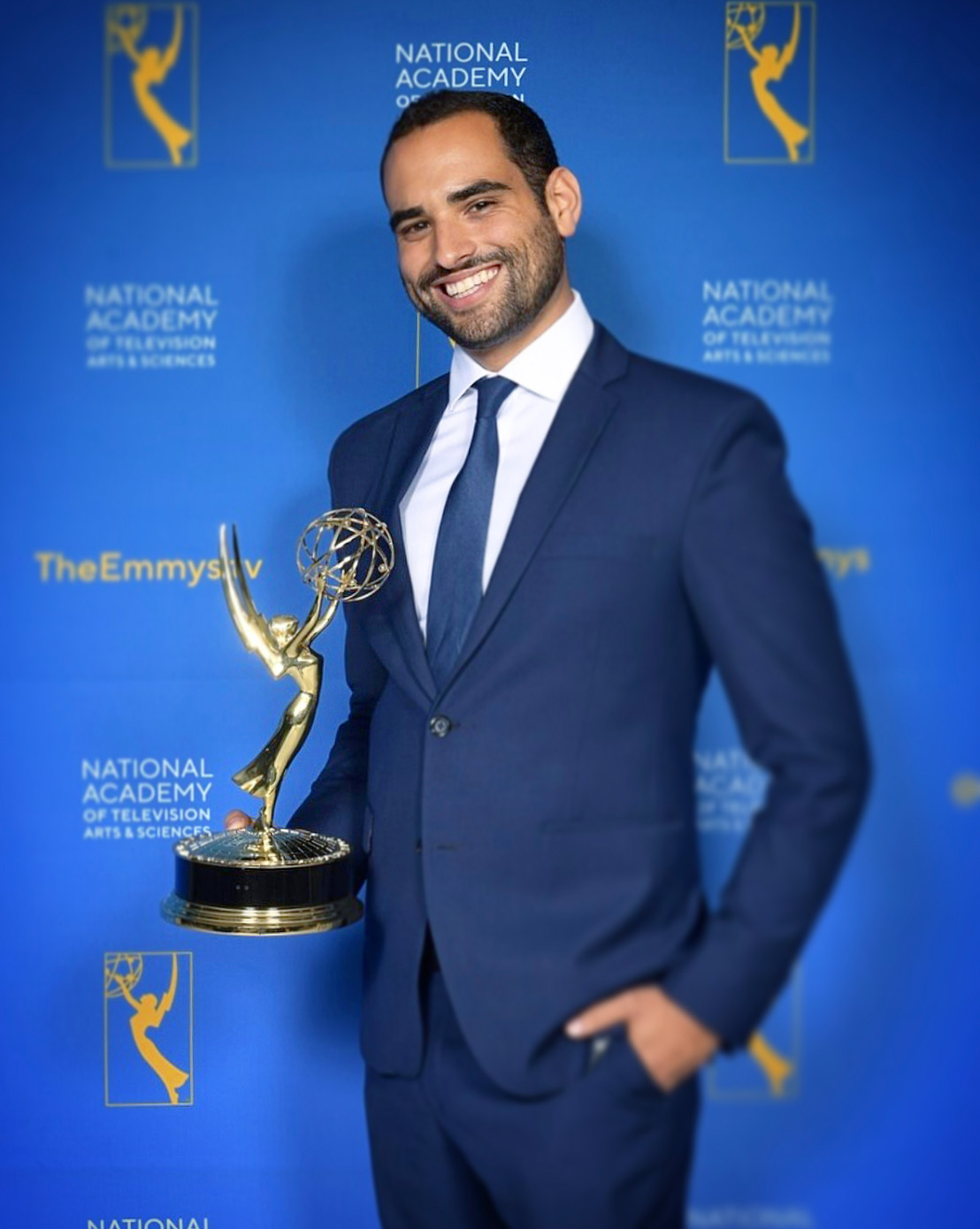 Smiling man in a blue suit holds an Emmy statuette against an awards backdrop.