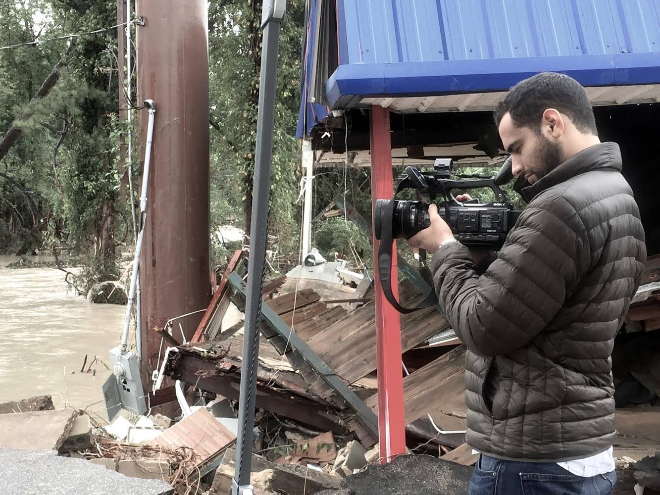 Man films floodwater and debris beside a damaged building.