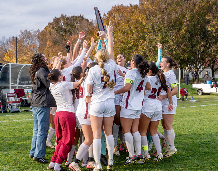 A sports team in white and red uniforms gathers in a huddle on a grass field, cheering as one member holds a trophy high in the air.