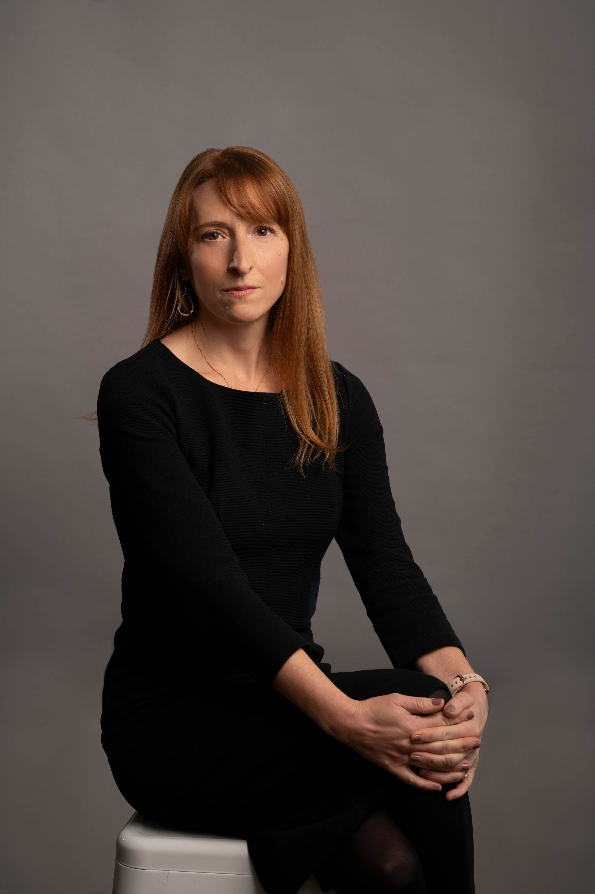 A person with shoulder-length reddish-brown hair and bangs looks directly forward against a solid gray backdrop, wearing a black top, a thin gold necklace, and a gold teardrop earring.