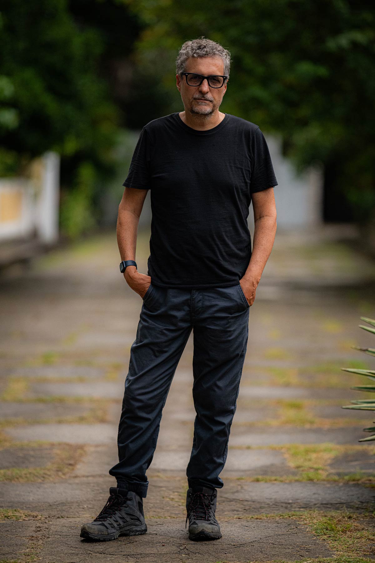 A person with short gray hair, facial hair, and black-framed glasses stands outdoors with hands in pockets, wearing a black t-shirt and dark pants against a blurred background of greenery.