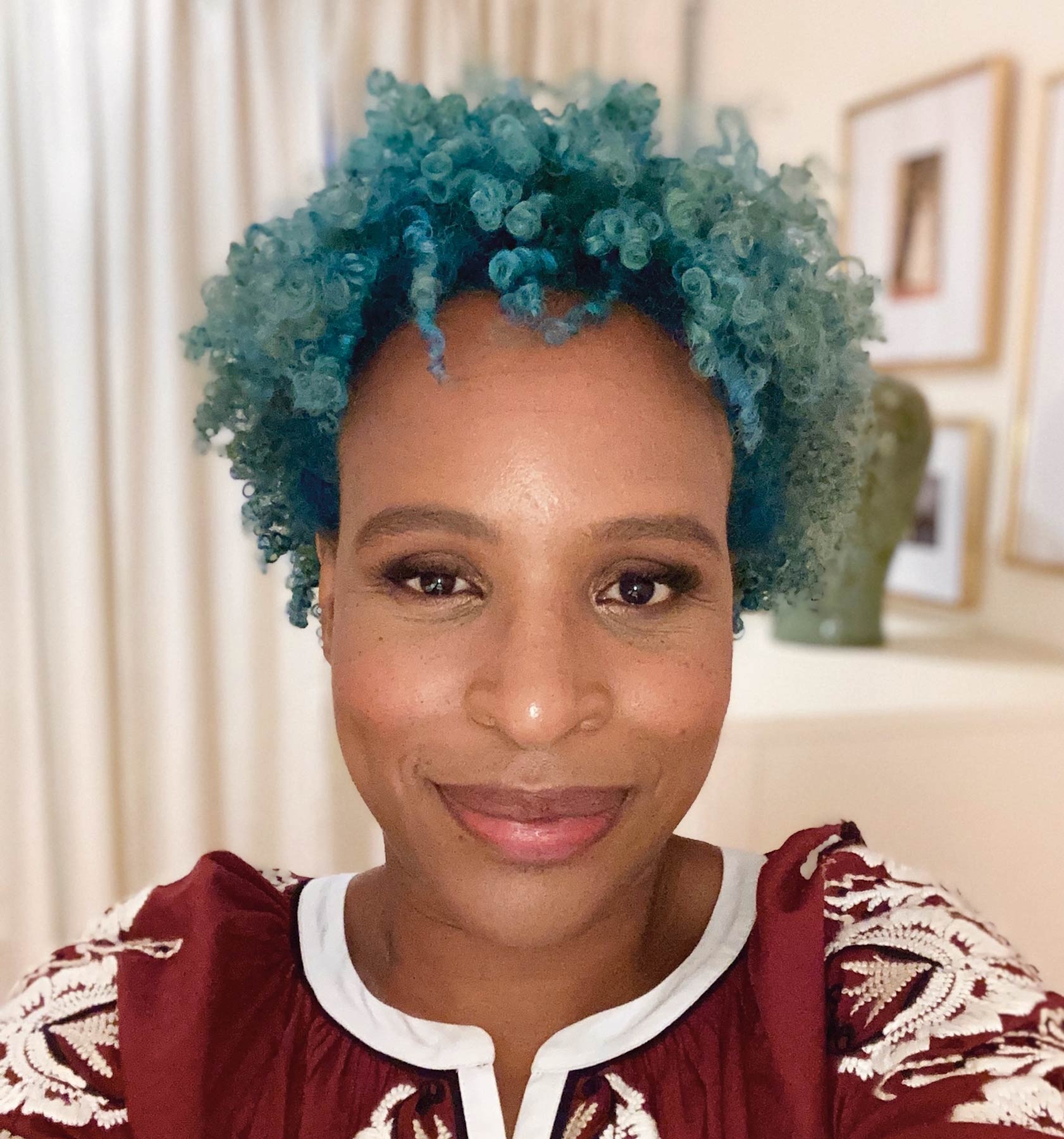 A person with short, bright blue curly hair smiles at the camera while wearing a maroon top with white embroidery. Light curtains and framed wall art are visible in the background.
