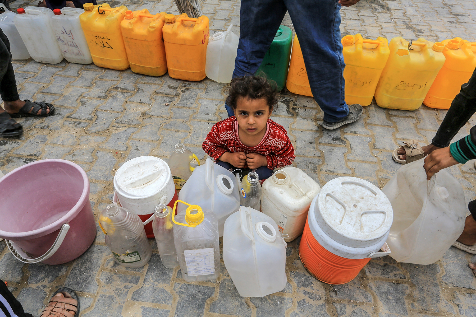 A small child sits on a cobblestone street amidst empty water jugs.