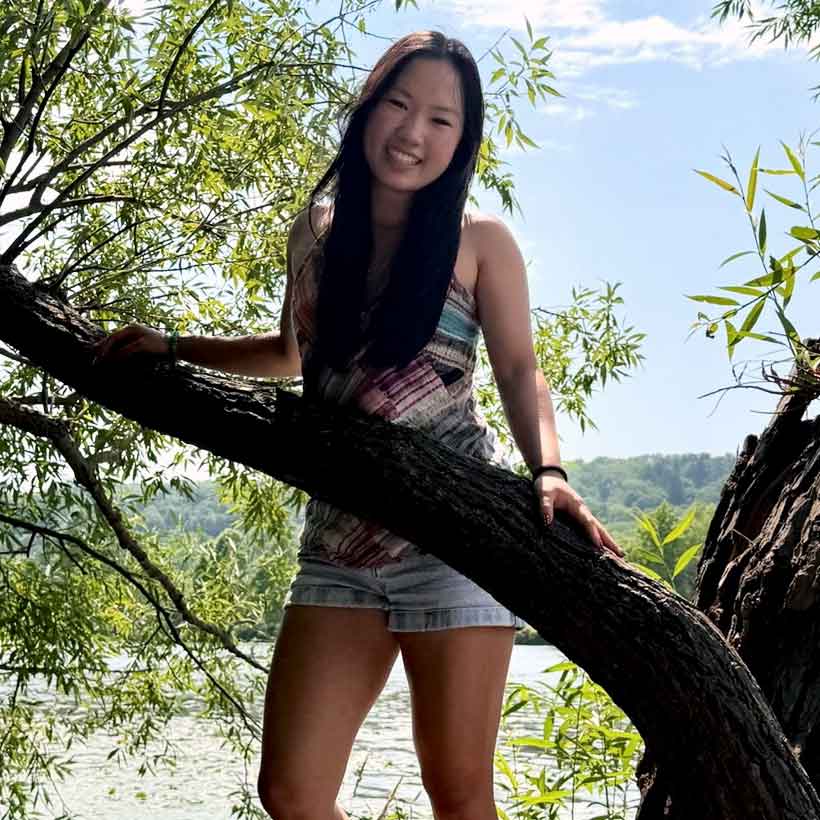 Photo of Sierra Chow standing behind a tree in front of a body of water.