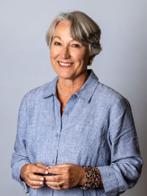 Susanne D. Lyons smiling, and is wearing a light blue linen shirt and a colorful beaded bracelet with with short, styled grey hair and her hands loosely clasped in front of her against a neutral grey background.