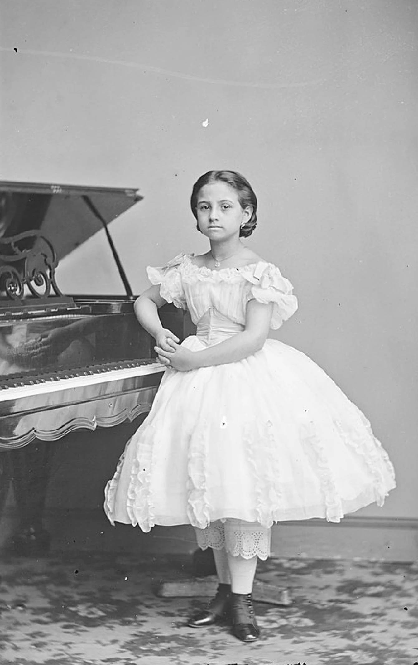 A person stands beside an open grand piano, wearing a full-skirted, off-the-shoulder white dress with lace-trimmed pantalettes and dark boots. The individual poses with hands clasped in front against a plain wall and patterned rug.