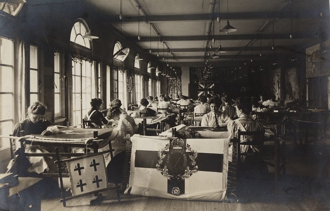 Early 20th century photograph of a large room where dozens of women sit working around needlework looms. Two flag-like needlework pictures are displayed in the foreground. 