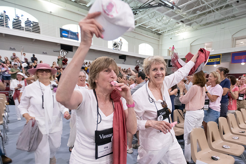 Reunion attendees wave a hat and scarf while a surrounding crowd applauds in a gymnasium.