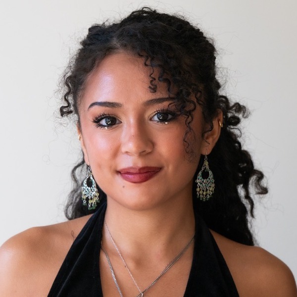 A portrait of Aiko Morales-Ishmon, a person with black curly hair, wearing a black halter top, earrings, and layered necklaces.