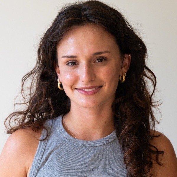 A portrait of Ari Gottsegen, a person with dark brown curly hair, wearing a gray sleeveless top and hoop earrings.