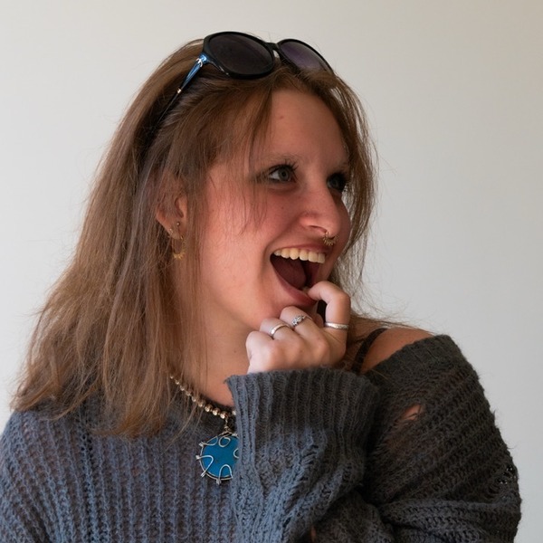 A portrait of Frieda Dangerfield, a person with light brown hair, wearing sunglasses on their head, a charcoal knit sweater, and necklaces.