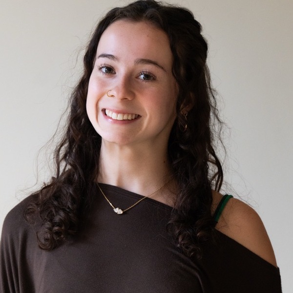 A portrait of Gwen Proctor, a person with dark brown curly hair, wearing a dark off-the-shoulder top and layered necklaces.