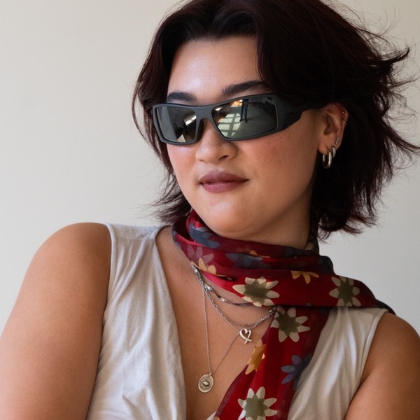 A portrait of Hana Colley, a person with dark auburn hair, wearing black sunglasses, a white sleeveless top, and a floral scarf.