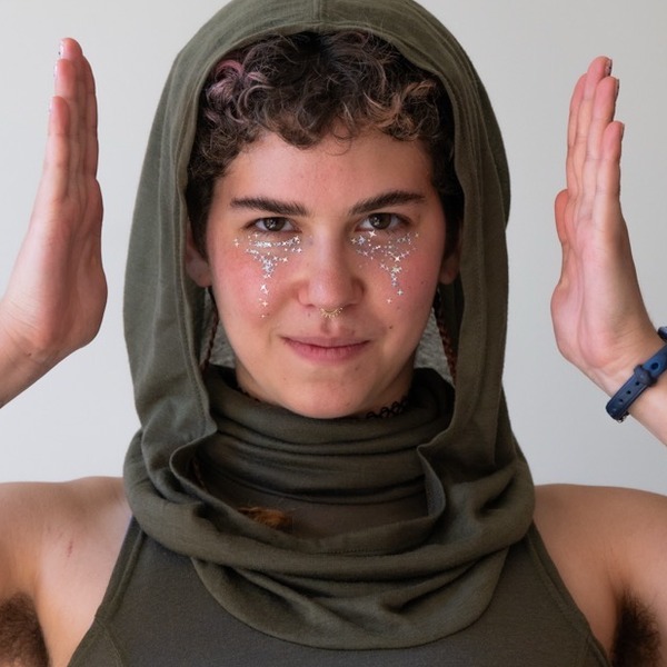 A portrait of Lila Ehrlich, a person with brown curly hair, wearing an olive hood and sleeveless top with both hands raised.