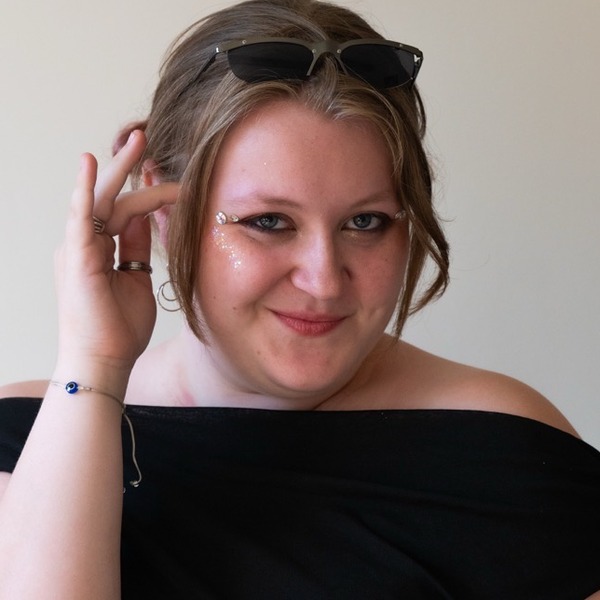 A portrait of Madi Quayle, a person with light brown hair, wearing sunglasses on their head and a black off-the-shoulder top.