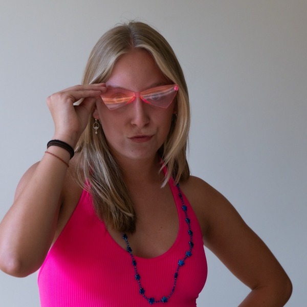 A portrait of Margo Walters, a person with blonde hair, wearing pink sunglasses, a bright pink sleeveless top, and a blue beaded necklace.