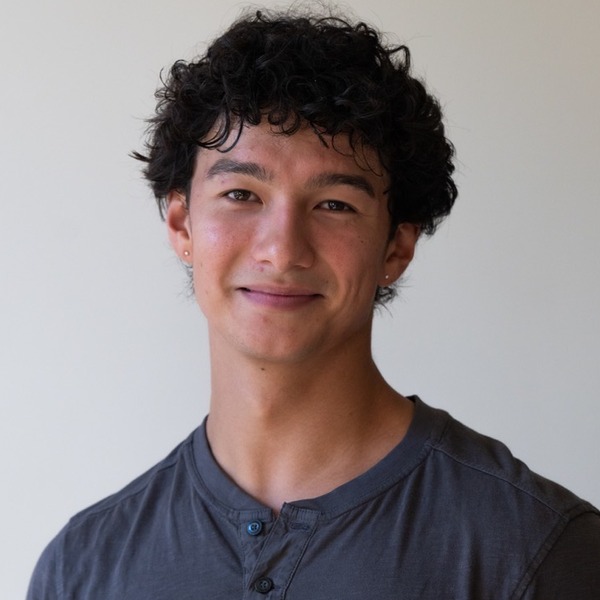 A portrait of Nikko Ramos, a person with black curly hair, wearing a gray long-sleeved henley shirt.